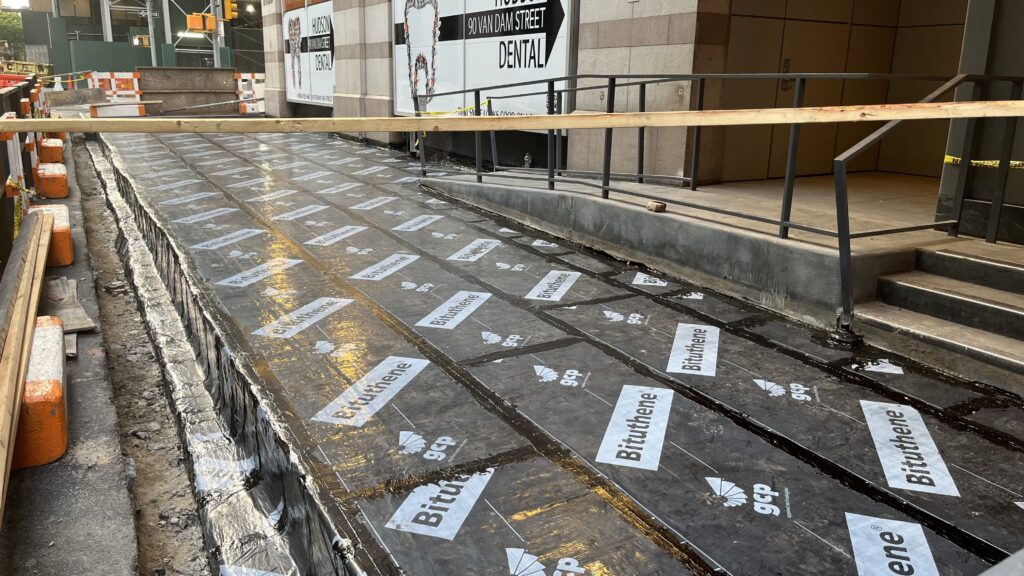 A Construction Site With a Sidewalk Covered in Black Waterproofing Membrane That Has “Bituthene” Logos Printed on It, Wooden Barriers on the Side, and Stairs Leading to a Dental Office in the Background.