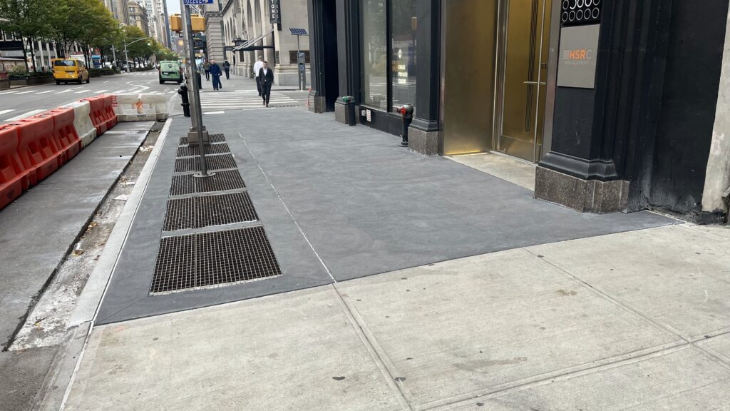 A City Street With a Sidewalk, Metal Grates, Red and White Plastic Barriers, and a Building With a Sign That Reads “Hsrc Management.”